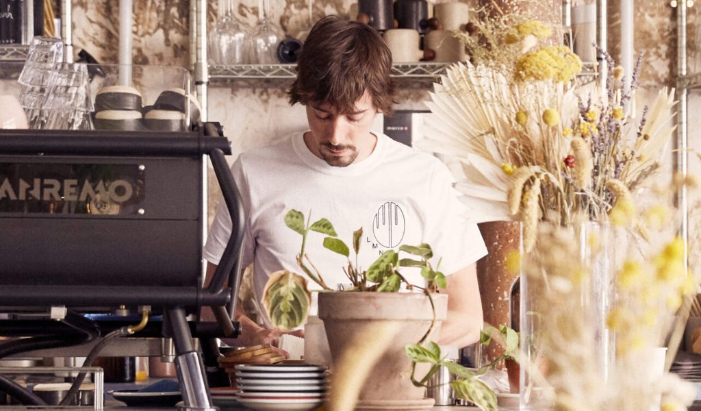 Personne préparant des boissons derrière un comptoir de coffee shop, entourée de plantes et de vaisselle