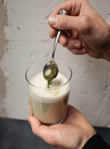 Mains tenant un verre de boisson lactée pendant qu’un liquide vert est versé à la cuillère
