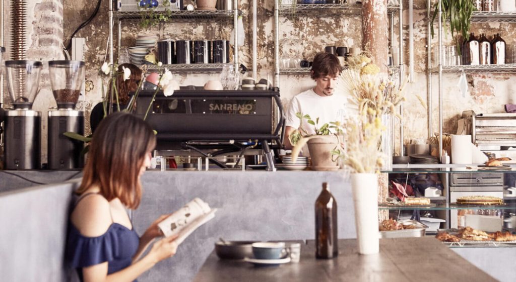 Personne lisant assise à une table pendant qu’un barista prépare des boissons derrière le comptoir d’un coffee shop